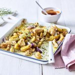 Op een witte tafel staat een witte ovenschaal met blauw randje. De ovenschotel is heerlijk gegaard uit de oven, aardappelstukjes, rode ui en kip. Ieders favoriet toch. Achteraan staat ook nog een potje met de homemade barbecuesaus. 