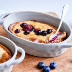 Op een houten plank staan twee ovenschaaltjes, deze zijn gevuld met heerlijke cheesecake uit de oven. In de cheesecake zitten blauwe bessen en braambessen, die zorgen voor een heerlijk zoet dessert!