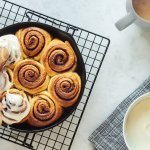 Op het werkblad staan de cinnamon rolls afgebakken in de bakvorm. Met een houten lepel smeren we de topping van roomkaas en suiker erover.