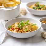 Op tafel staan twee bordjes uitgeschept met de oranje curry met stukjes aardappel, pompoen en visrolletjes. Het gerechtje is afgewerkt met gebakken kikkererwten en koriander. 