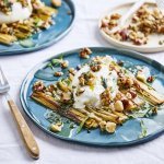 Twee blauwe borden staan op tafel met een wit tafelkleed. Op het bord liggen reepjes geroosterde prei met hierop burrata, notencrumble en dragonolie. Nog enkele blaadjes dragon voor extra groen. Prachtig om naar te kijken en heerlijk om te proeven!