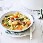 Er staat een soepbordje op tafel, boordevol lekkers. Je ziet de gnocchi, stukjes kerstomaat, spinazie en een gebakken chipolata. De soepkom zelf is wit en staat rechtsbovenaan in beeld. 