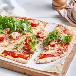 Op een houten snijplank ligt een aangesneden pizza. Hij is belegd met tomatensaus, ham, appelplakjes en Passendale kaas. We werken hem af met rucola en pijnboompitjes.