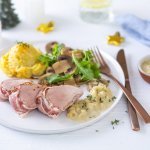 Op tafel staat een geserveerd bord met stukjes varkensvlees, gegratineerde knolselderpuree en rucola met gebakken champignons.