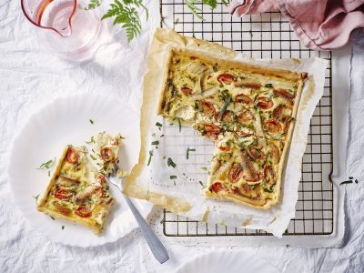 Vers gebakken uit de oven staat de quiche met asperges, tomaten en gerookte paling te blinken op tafel. Er werd al een stukje aangesneden en op een bordje gepresenteerd. Roze linnen servet en waterkaraf zorgen voor de sfeer in de foto. 