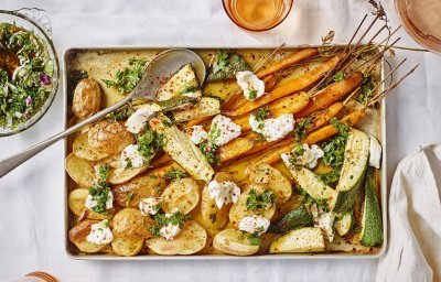 Ovenplaat met gegrilde groentjes komt net uit de oven en staat al op tafel. Hij heeft warme kleuren van de gouden aardappelen naar de oranje wortelen. Hij is afgewerkt met toefjes geitenkaas en verse chimichurri.