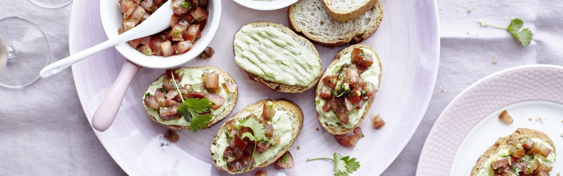 Bruschetta met een yoghurtdip en tomaten Bruschetta's liggen op een pastelroos bord, samen met een wit potje waarin de yoghurtdip zit en een wit potje waarin stukjes versgesneden tomaat en kroiander zit. De gasten smeren dus zelf hun bruschetta, waardoor de gastvrouw of -heer extra tijd aan tafel krijgt.